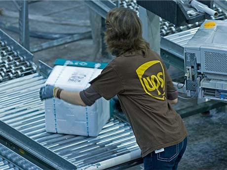 A UPS employee wearing the iconic brown and yellow uniform puts a square, white box on conveyer belt in Georgia.