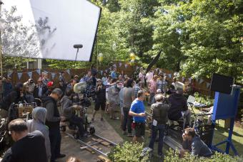 OverviA large group of people behind the scenes of filming outdoors in a backyard with cast on set