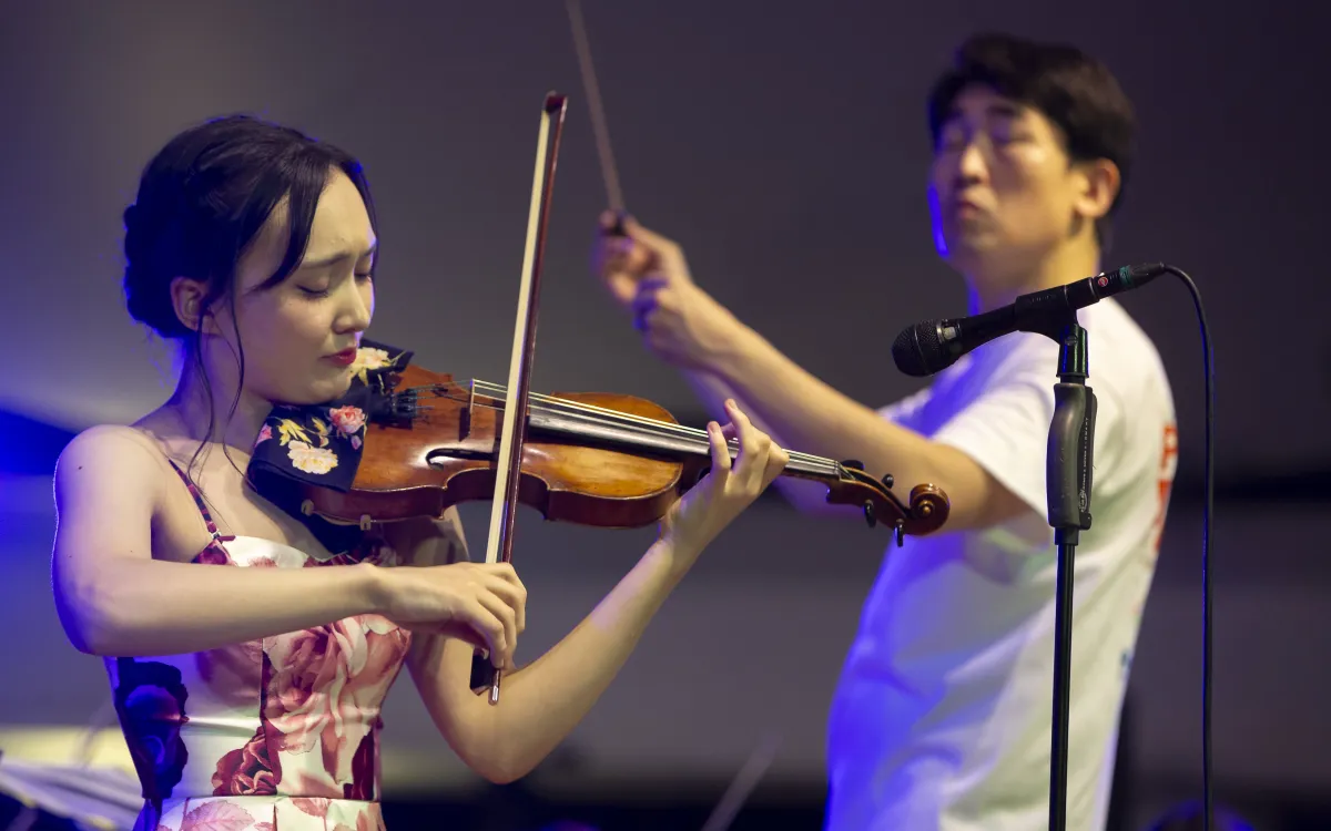 young woman playin violin on stage with a conductor in the background