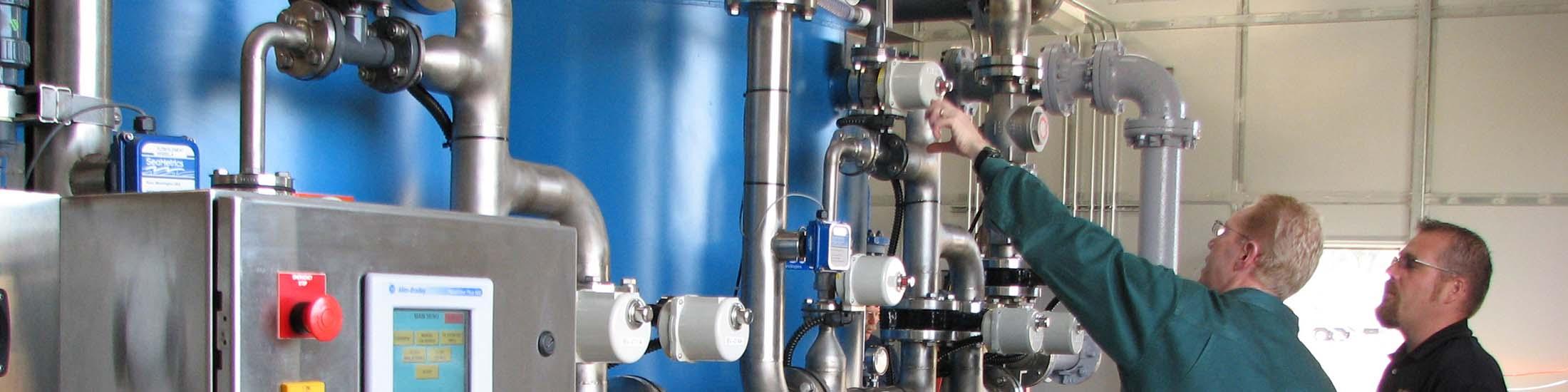 Two people wearing glasses look at the meters on a complex system of pipes at a manufacturing facility in Georgia.