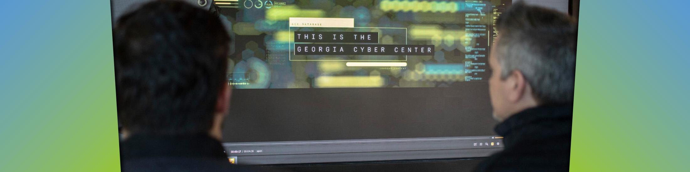 Two people wearing dark-colored turtle necks look at a screen with blue and green blurred images and says "This is the Georgia Cyber Center"