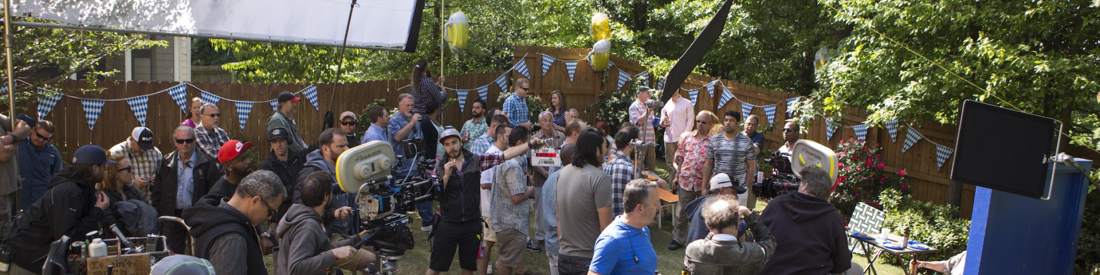 Overview photo of large group of people behind the scenes of filming outdoors in a backyard with cast on set