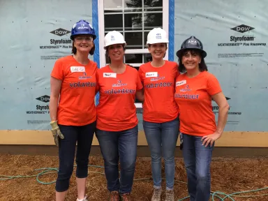 Four women in orange shirts and hard hats stand in front of a building under construction