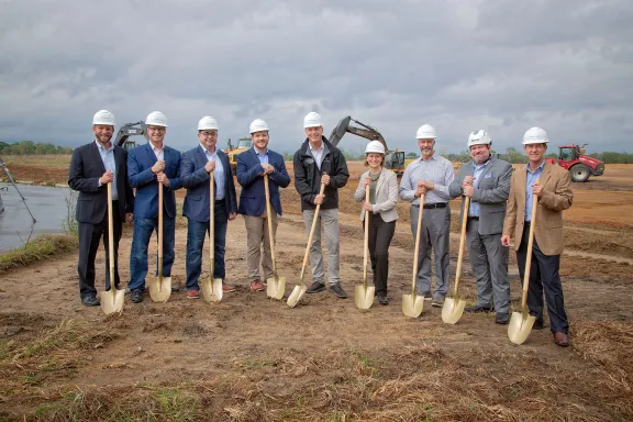Nine people in hardhats stand with shovels at a construction site with heavy equipment in the background